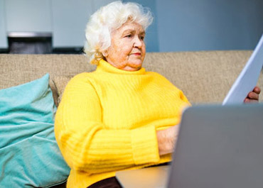 Senior woman in yellow sweater looking at documents, with a laptop, symbolizing vulnerable victims.