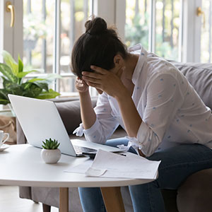 Person with head in hands, looking at laptop and papers, symbolizing financial stress.