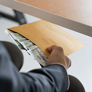 Hands exchanging a brown envelope filled with money under a desk, symbolizing embezzlement.