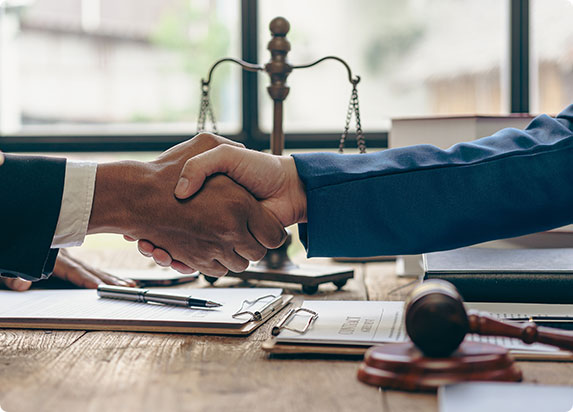 Two individuals shake hands across a table with legal documents, a pen, a gavel, and scales of justice, symbolizing a legal agreement or consultation.