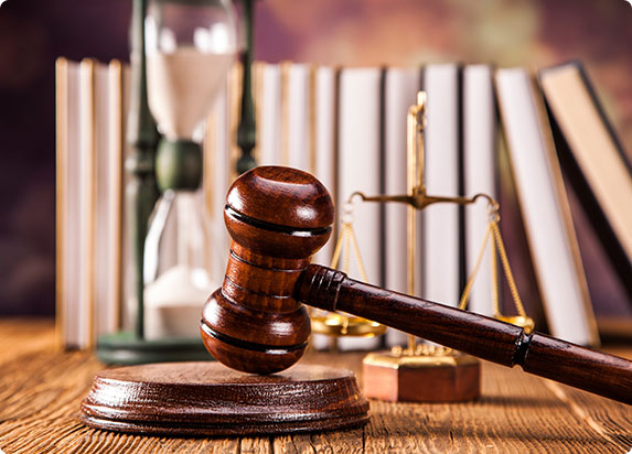 A close-up of a wooden gavel resting on a sound block, with an hourglass and legal books blurred in the background.