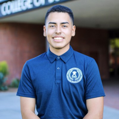Smiling male student in a dark blue polo shirt standing outside a building represents the Community Leaders Scholarship.